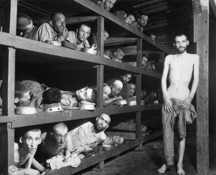 Osvobození vězni v koncentračním táboře Buchenwald. V prostřední řadě se jako sedmý zleva (vedle sloupku) do objektivu dívá Elie Wiesel, který za 13 let jako první veřejně použije termín „holokaust“. Foto: US Army, H. Miller, Wikimedia Commons