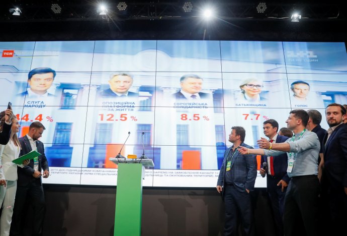 Ukrajinský prezident Volodymyr Zelenskyj reaguje na průběžné výsledky parlamentních voleb na velkoplošné obrazovce v centrále jeho strany Služebník lidu. Její výsledek je zcela vlevo, 44,4 % s fotografií lídra volebního strany Dmytra Razumkova. Kyjev, 21. 7. 2019. Foto: Valentyn Ogirenko, Reuters