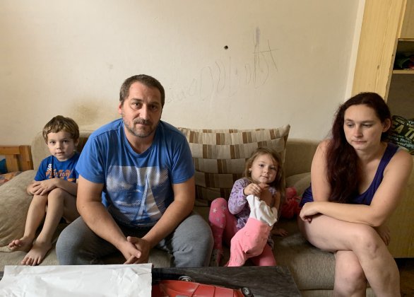 Pohovka, na které manželé Břízovi a jejich nejmenší děti sedí, je zároveň i jejich manželskou postelí. Dcera a syn s nimi spí v jedné místnosti, ostatní děti spí ve vedlejším, průchozím pokoji na palandách. Foto: Jana Ciglerová, Deník N