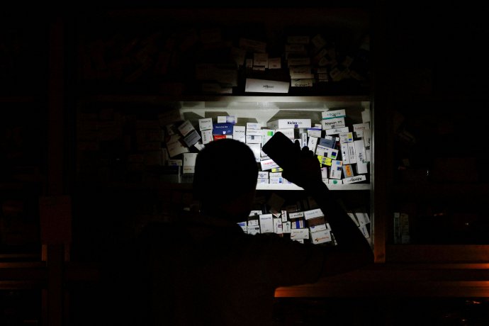 Prodavač v lékárně si během výpadku elektřiny kvůli nedostatku plynu svítí na léky v regálu telefonem. Foto: Muhammad Hossain, Reuters